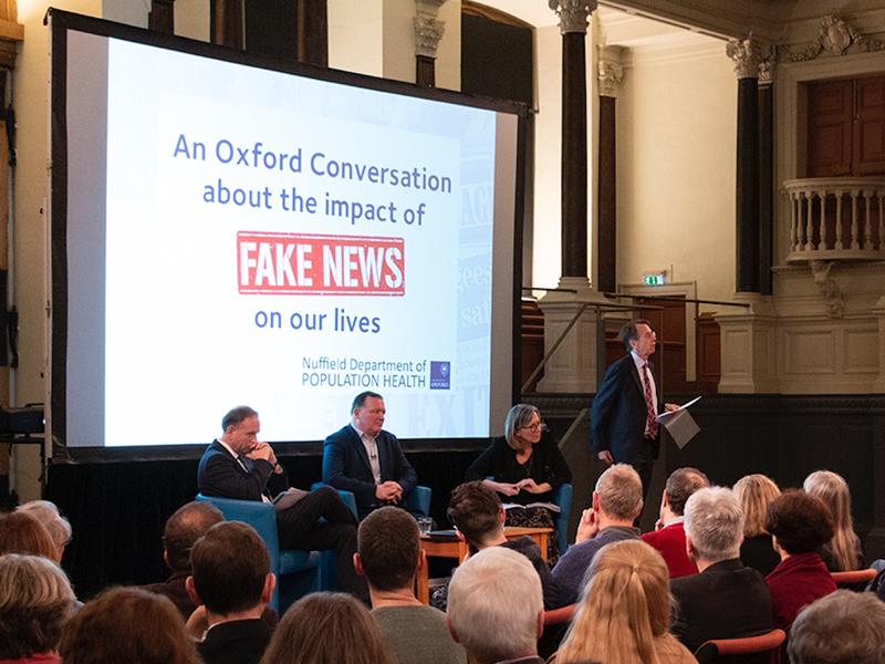 Fake news discussion - Credit: Nuffield Department of Population Health/Graham Bagley A view from within the audience of a stage with four speakers, and on the screen is a slide saying 'An Oxford Conversation about the impact of Fake News on our lives'